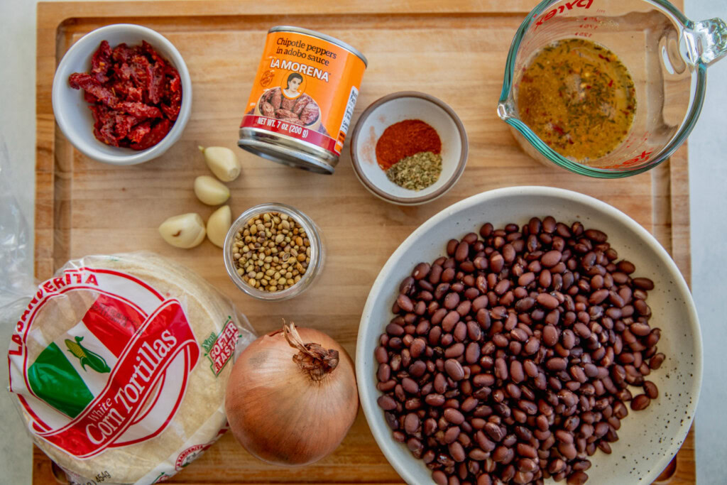Cutting board filled with sun-dried tomatoes, onion, chipotle peppers, veggie broth, spices, garlic, black beans, and tortillas.