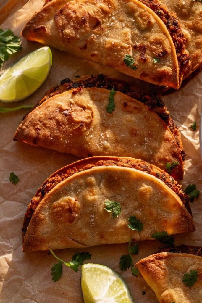 Close up of a bunch of black bean tacos on a work surface surrounded by lime wedges and cilantro.