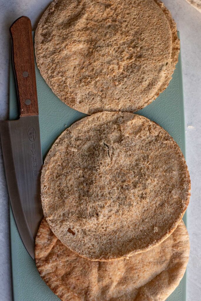 Cutting the pita in half lengthwise to create 2 equal rounds on a cutting board.