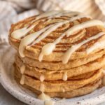 Vegan cinnamon roll pancakes stacked on top of each other and drizzled with maple cream cheese glaze.