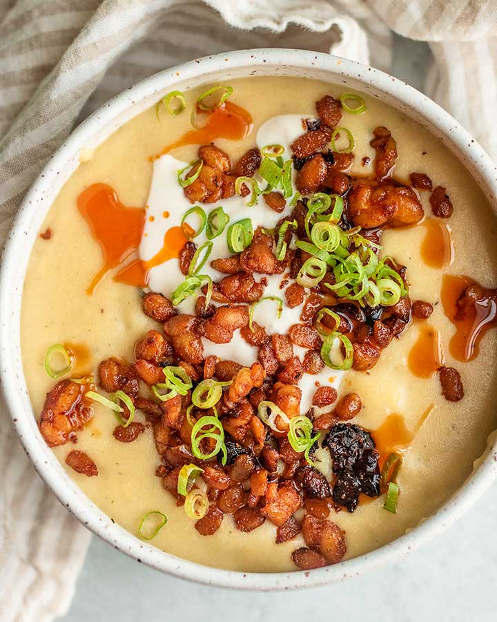Close up view of a bowl of potato soup topped with vegan sour cream, tempeh crumbles, and scallions.