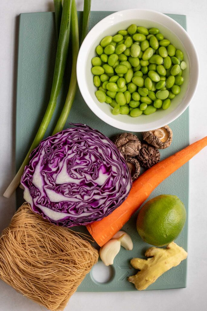 Cutting board with some carrot, cabbage, a bowl of edamame, mushrooms, lime, ginger, garlic, and scallions.