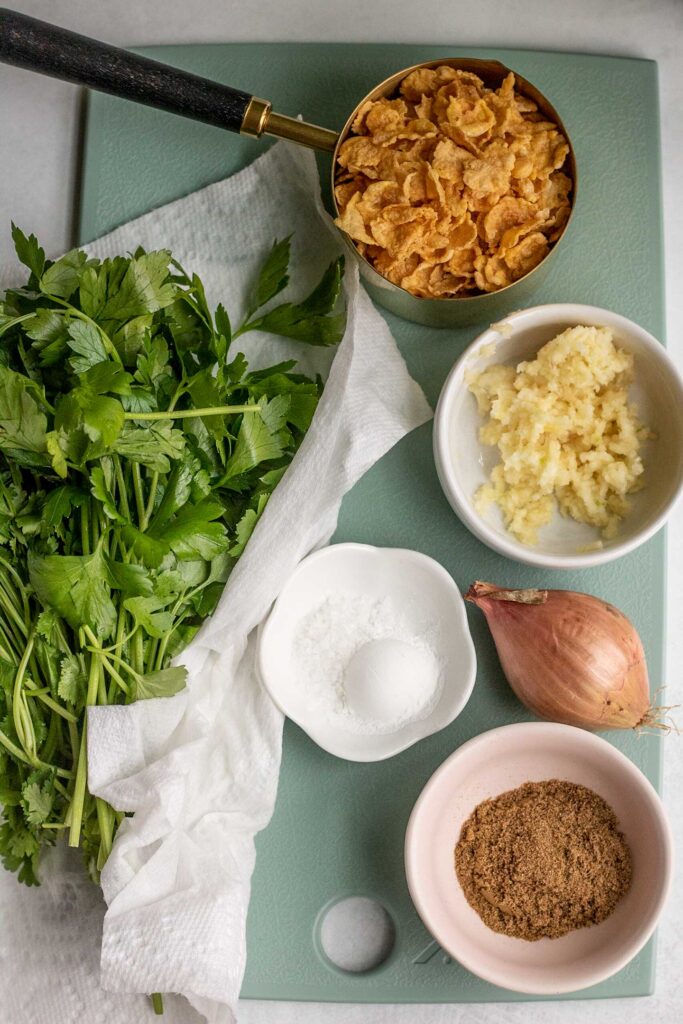Cutting board topped with cornflakes, minced garlic, baking powder, shallot, spices, fresh herbs, and chickpeas.