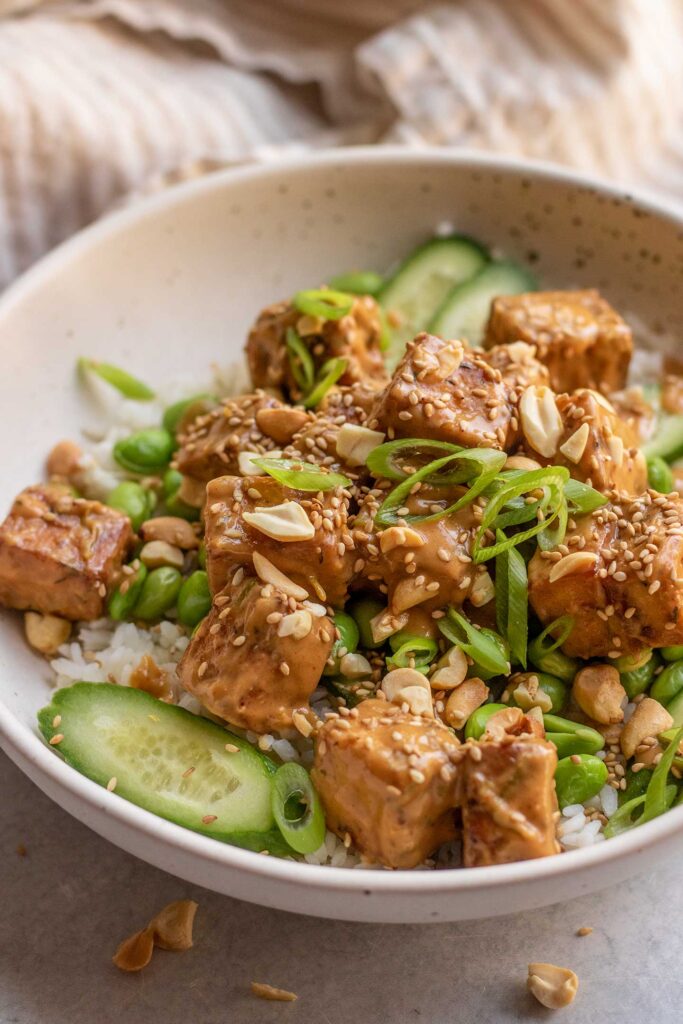 Peanut tofu topped over a bowl of rice, edamame and cucumber then topped with scallions and peanuts.