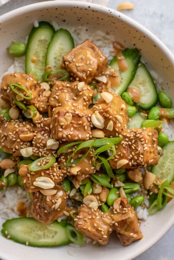 Close up view of a bowl of tofu coated in peanut sauce served with rice, edamame, and cucumbers.