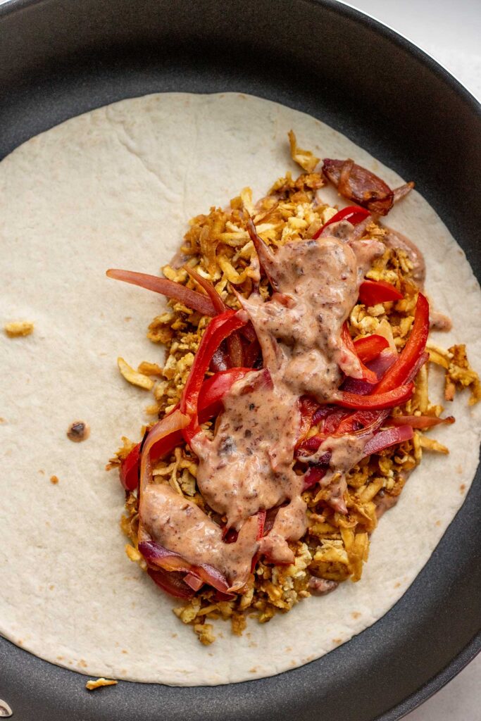 Large tortilla with half of it filled with chipotle mayo sauce, shredded tofu, and peppers and onions.