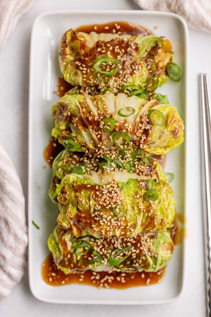 Top down view of a rectangular white plate topped with cabbage rolls, sticky sauce, sesame seeds and scallions.
