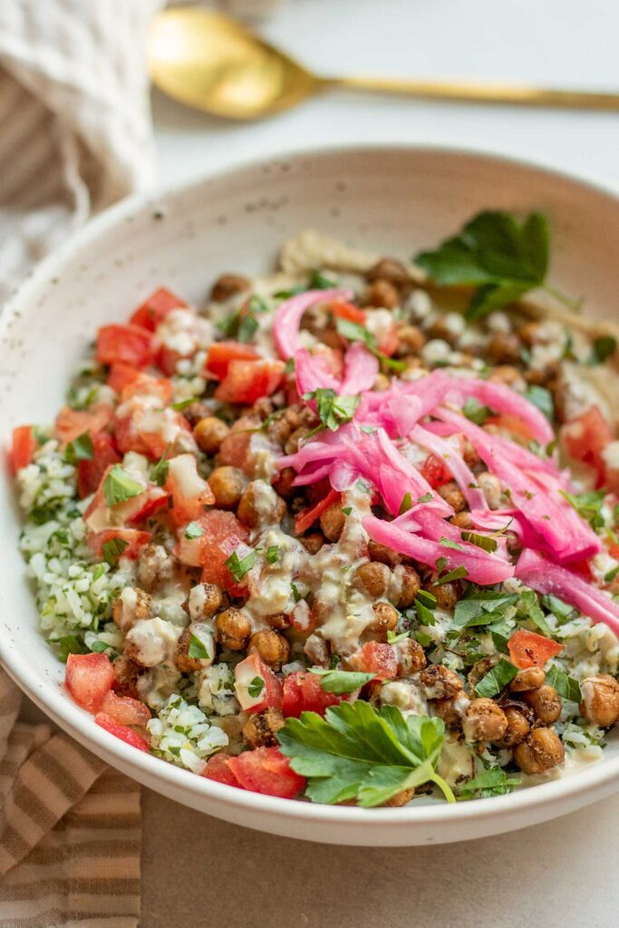 Side view of a white bowl filled with hummus and topped with herby mixed rice, roasted chickpeas, pickled onions and a hummus dressing.