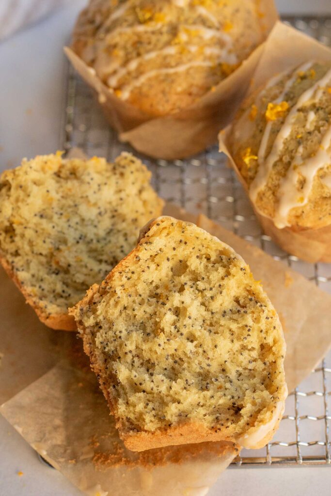 One orange poppy seed muffin broken in half and laying next to two full muffins on a grate.