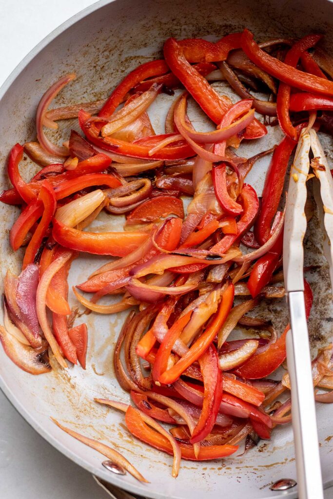 Sauteing red bell peppers strips and onion slices in a pan with tongs.