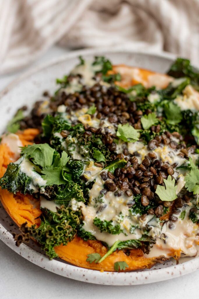 Side view of a plate stuffed sweet potatoes topped with crispy lentils, kale and a creamy tahini dressing on top.