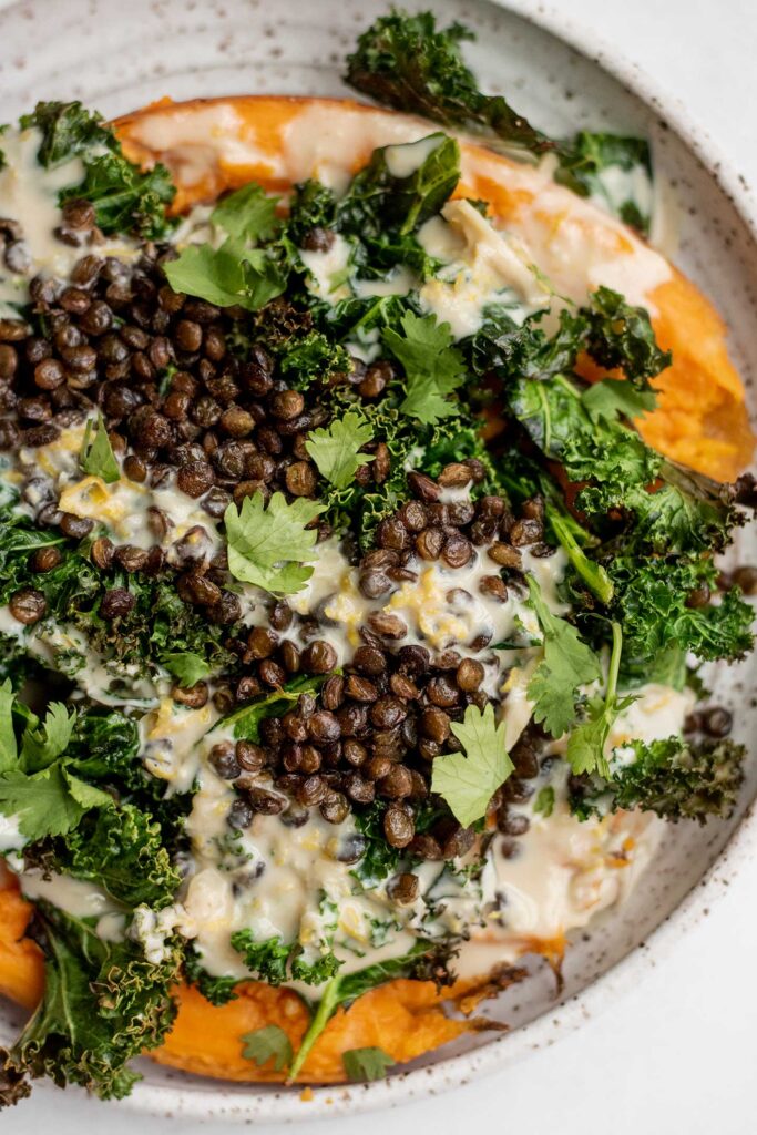White plate topped with a mashed roasted sweet potato stuffed with kale and crispy lentils then drizzled with a tahini sauce.