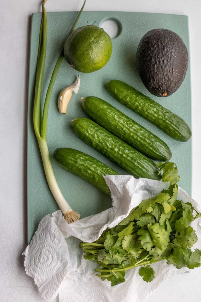 Persian cucumbers, avocado, scallion, lime, cilantro and garlic clove on a blue cutting board.