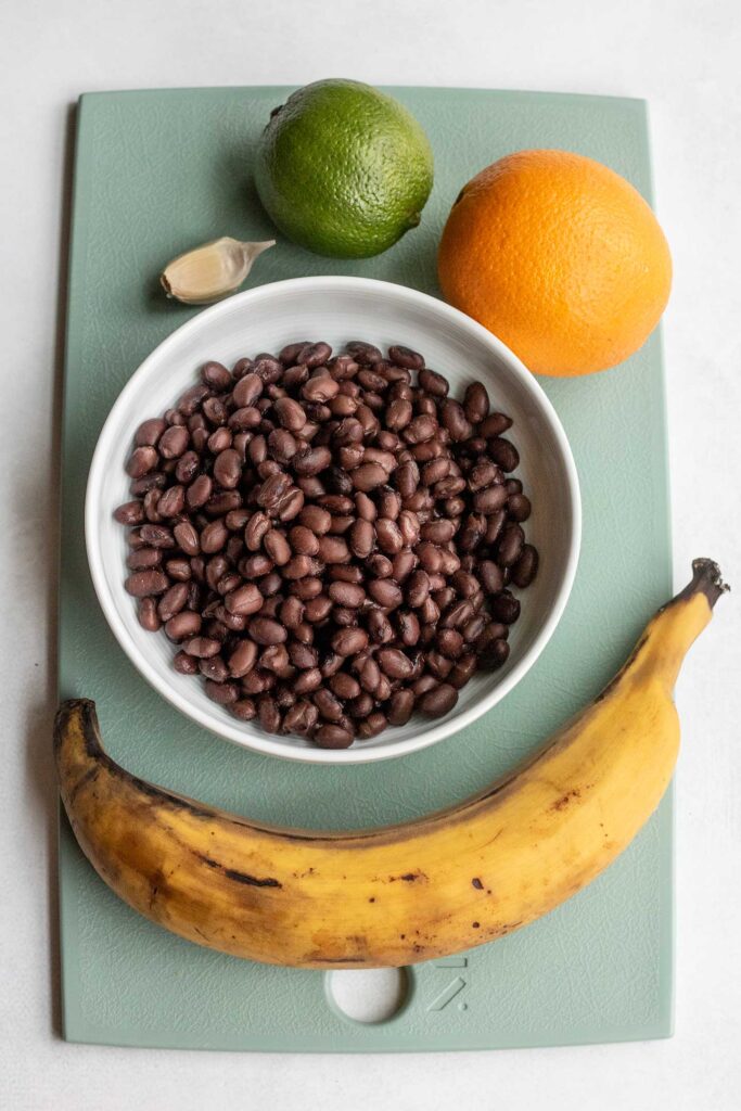 Cutting board with a bowl of black beans, plantains, orange, lime and garlic on it.