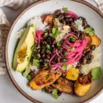 Top down view of a white bowl filled with crispy chunks of rice, baked plantain slices, black beans, avocado quarter and red pickled onions.