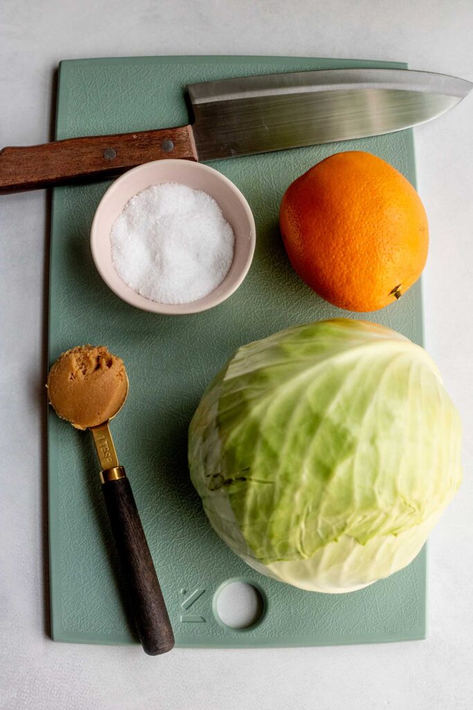 Cutting board with cabbage, orange, pinch bowl filled with salt, a tablespoon of miso paste and a knife on top.
