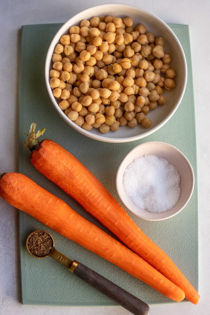 Bowl of chickpeas, two large carrots, salt and za'atar placed on a light blue cutting board.