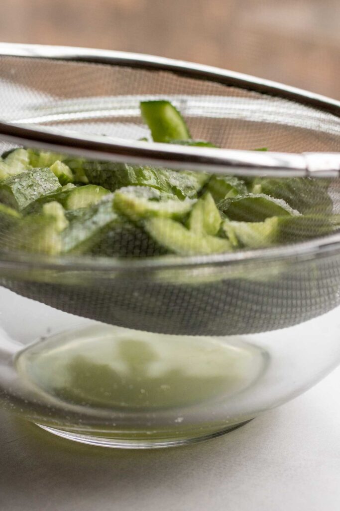 A mesh strainer filled with salted smashed cucumbers placed over a bowl to collect the strained water.