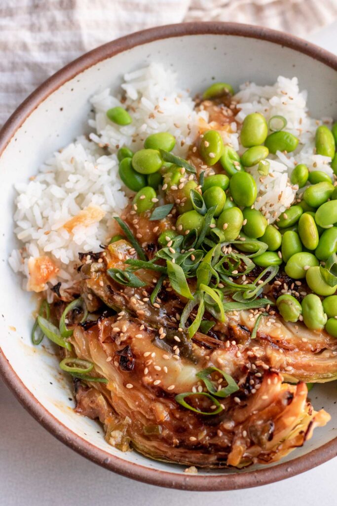 Close up of roasted cabbaged covered in orange miso sauce served with edamame, white rice, scallions and sesame seeds on top.