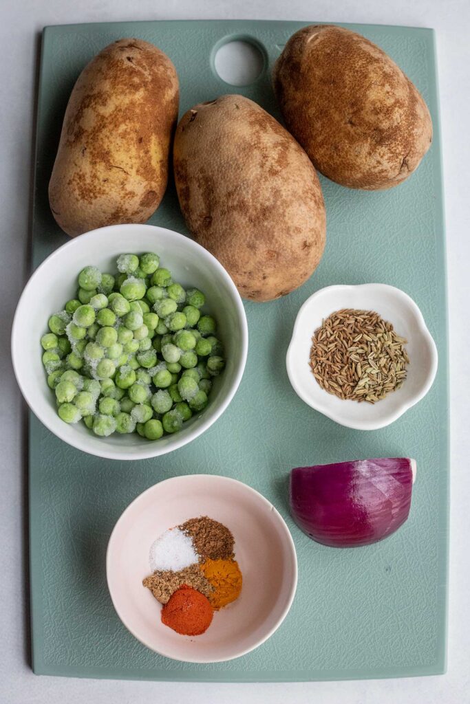 Cutting board with bowl of frozen peas, spices, red onion, and russet potatoes on top.