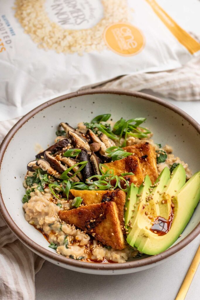 A side view of a bowl of savory oats topped with sliced mushrooms, pan-fried tofu, avocado and scallions.