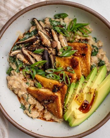 Top down view of a white bowl filled with miso oatmeal then topped with tofu triangles, avocado slices, sliced mushrooms and scallions.