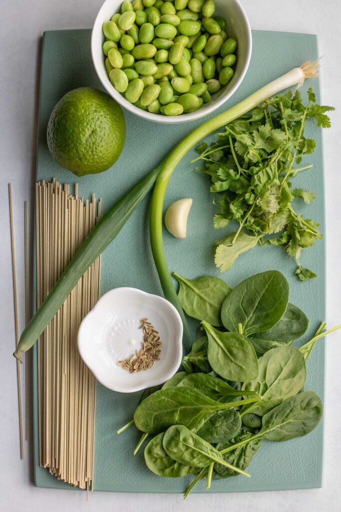 Cutting board topped with lime, edamame, scallion, cilantro, spinach, a small bowl of fennel seeds, and a garlic clove.