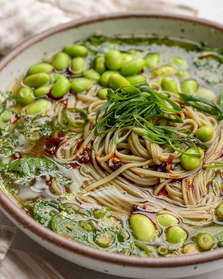 White bowl filled with a soy miso based broth and topped with soba noodles, edamame and scallions.