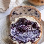 Blueberry bagel cut in half on a plate with one half smeared with cream cheese and blueberry jam.