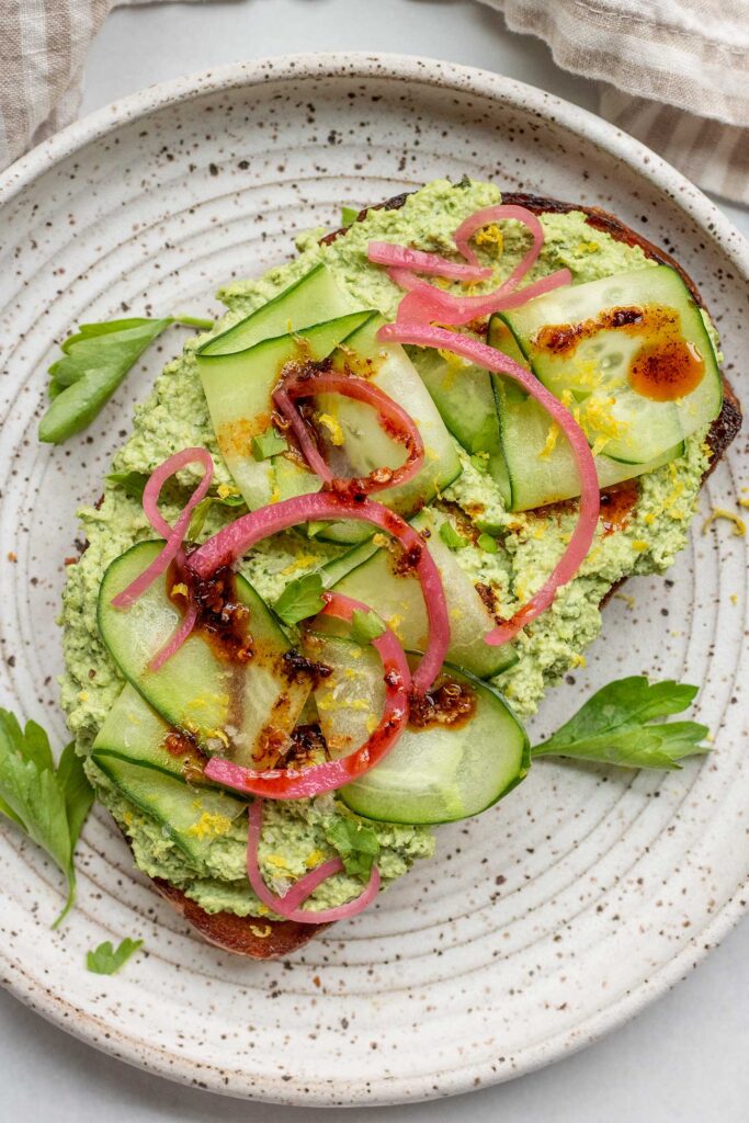 Toast on a white plate loaded with edamame spread, cucumber slices, pickled onions, chili oil, and parsley.