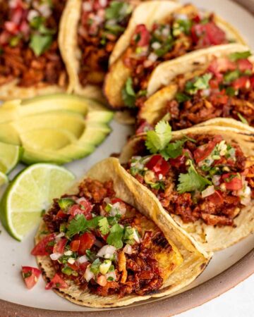 Close up of an arc of tacos stuffed with shredded tofu, pico de gallo and served with avocado slices and lime wedges on the side.