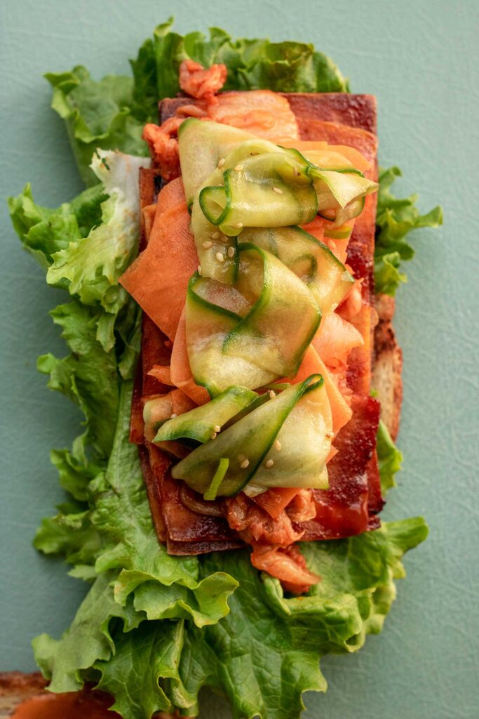 Slices of sandwich bread topped with lettuce, baked tofu slices, pickled carrots and cucumber, and kimchi.