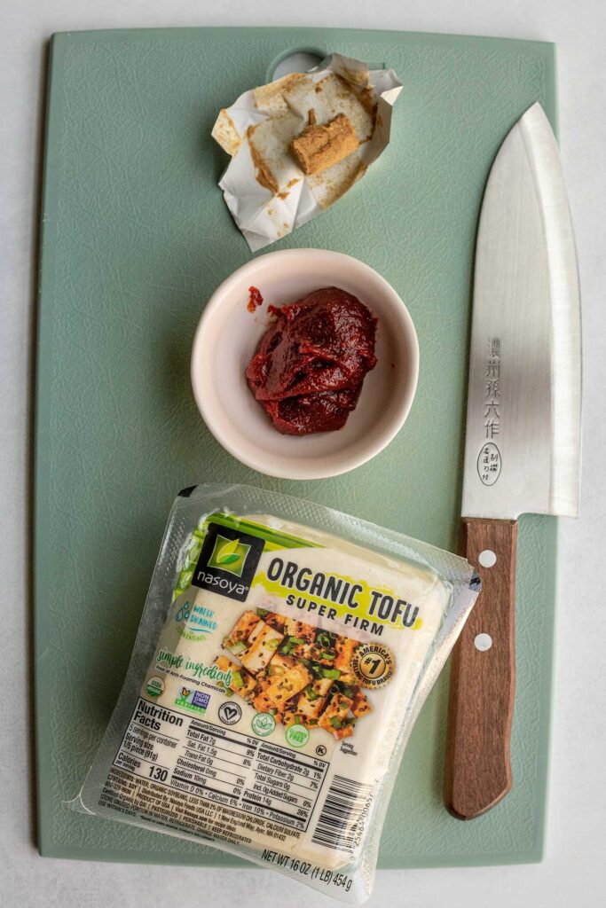 Tofu, gochujang, vegan bouillon cube and a knife placed on a cutting board.