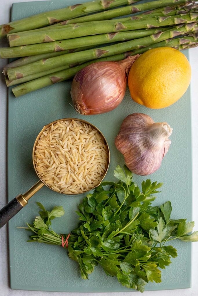Orzo, parsley, shallots, garlic, lemon and asparagus on a blue cutting board.