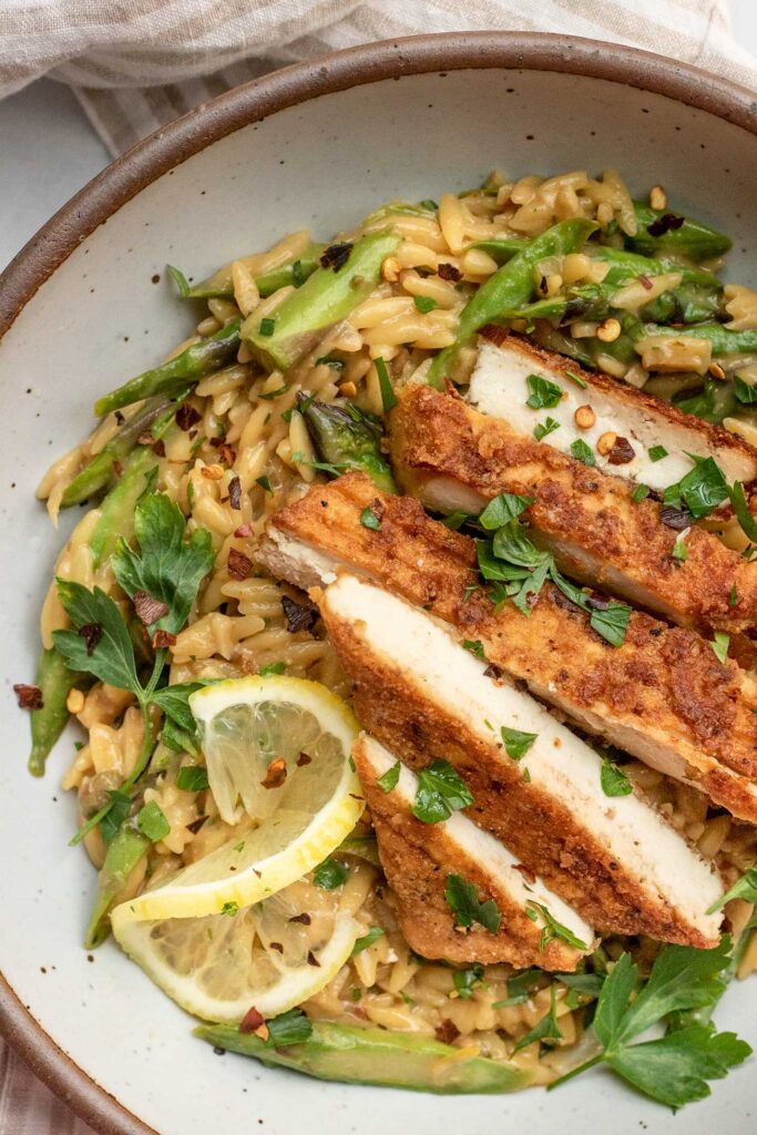 Close up of a bowl of orzo mixed with asparagus and topped with sliced tofu cutlet.