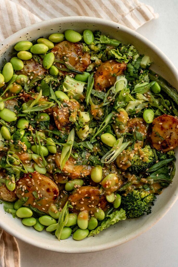 Bowl of roasted baby potatoes, broccoli, sugar snap peas, edamame, topped with tahini sauce, sesame seeds and scallions.
