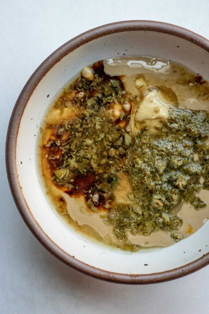 Tahini, capers, nutritional yeast, garlic and lemon juice in a small white bowl.