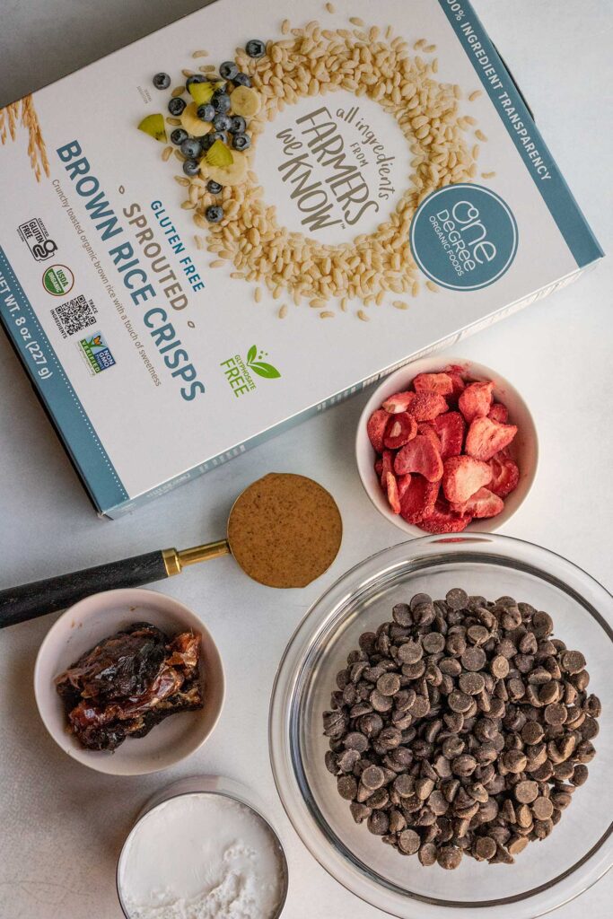 Box of cereal, bowl of chocolate chips, freeze-dried strawberries, a measuring cup with almond butter, and bowl of dates on a cooking surface.