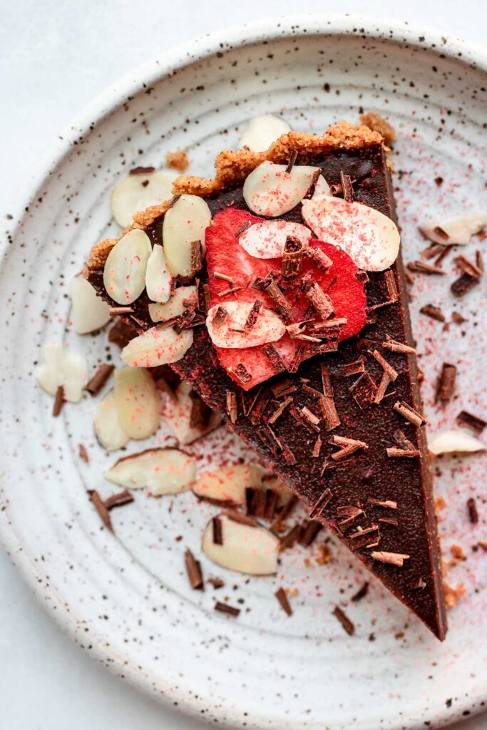 Slice of chocolate strawberry tart topped with extra almond slices, strawberry slices and shaved chocolate on a serving plate.