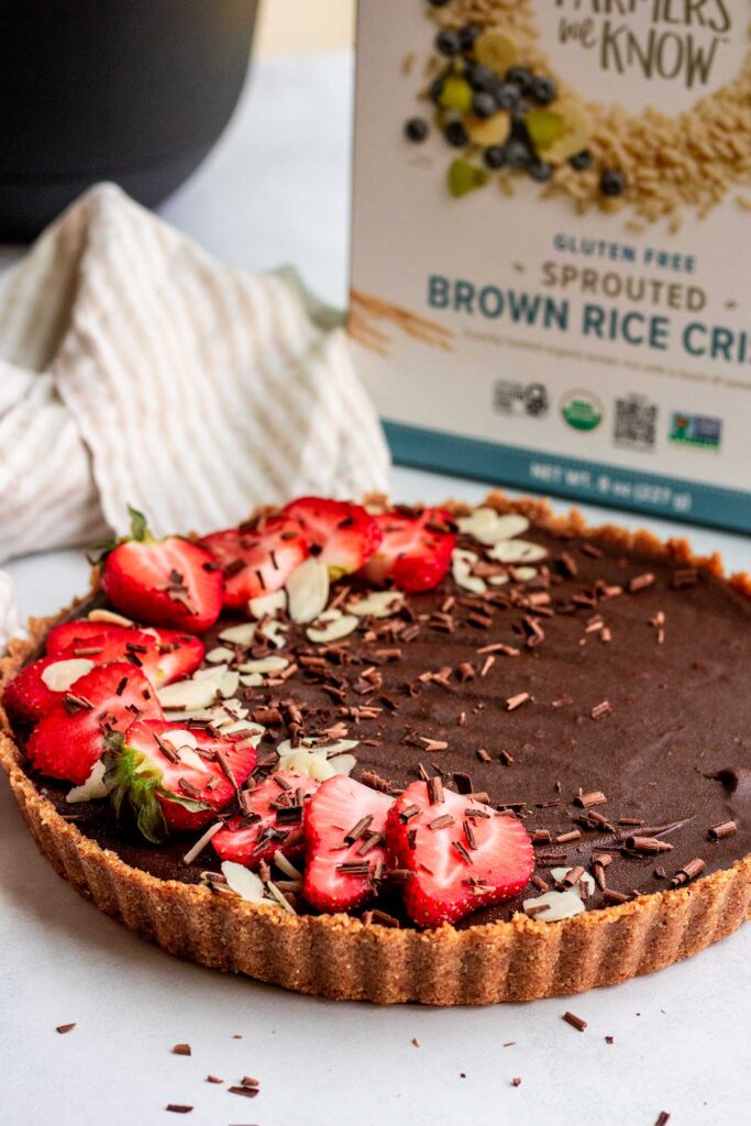 Chocolate strawberry tart topped with fresh sliced strawberries, shaved chocolate and almond slices with a box of cereal in the background.