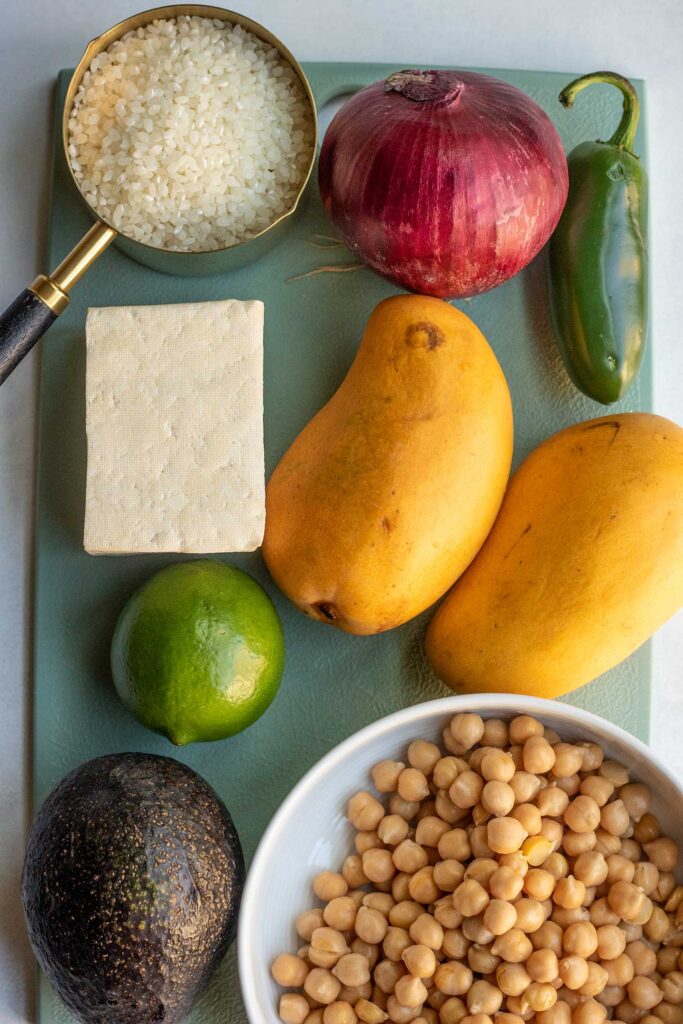 Cutting board topped with tofu, rice, onion, cucumber,mango, chickpeas, lime and avocado.