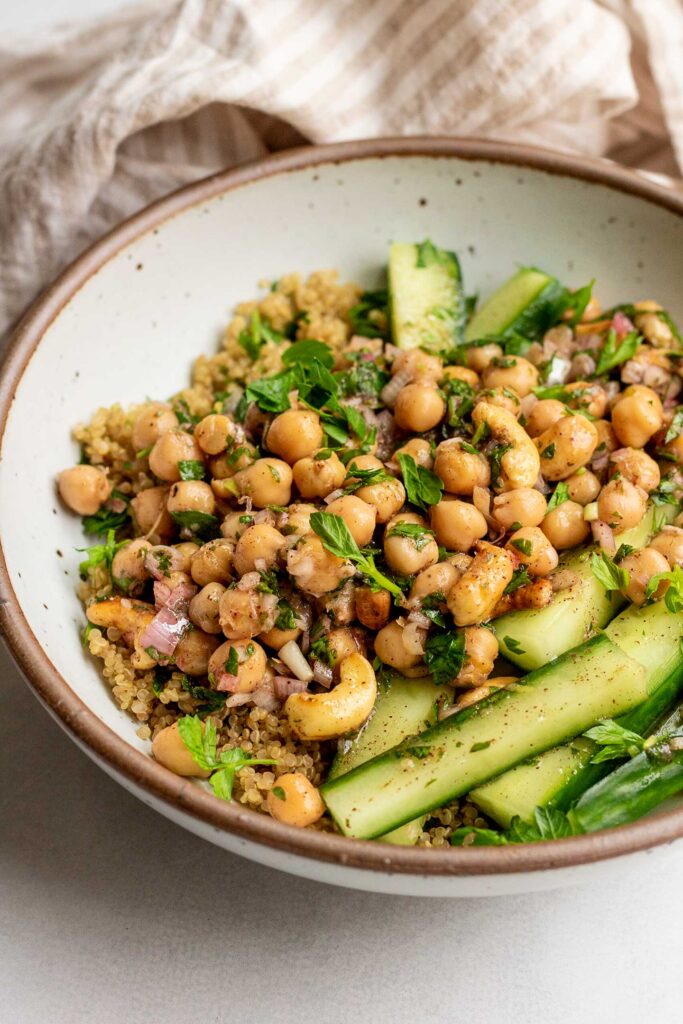 Side view of a bowl of chickpeas, cashews mixed together in a marinade served over quinoa and cucumber spears.