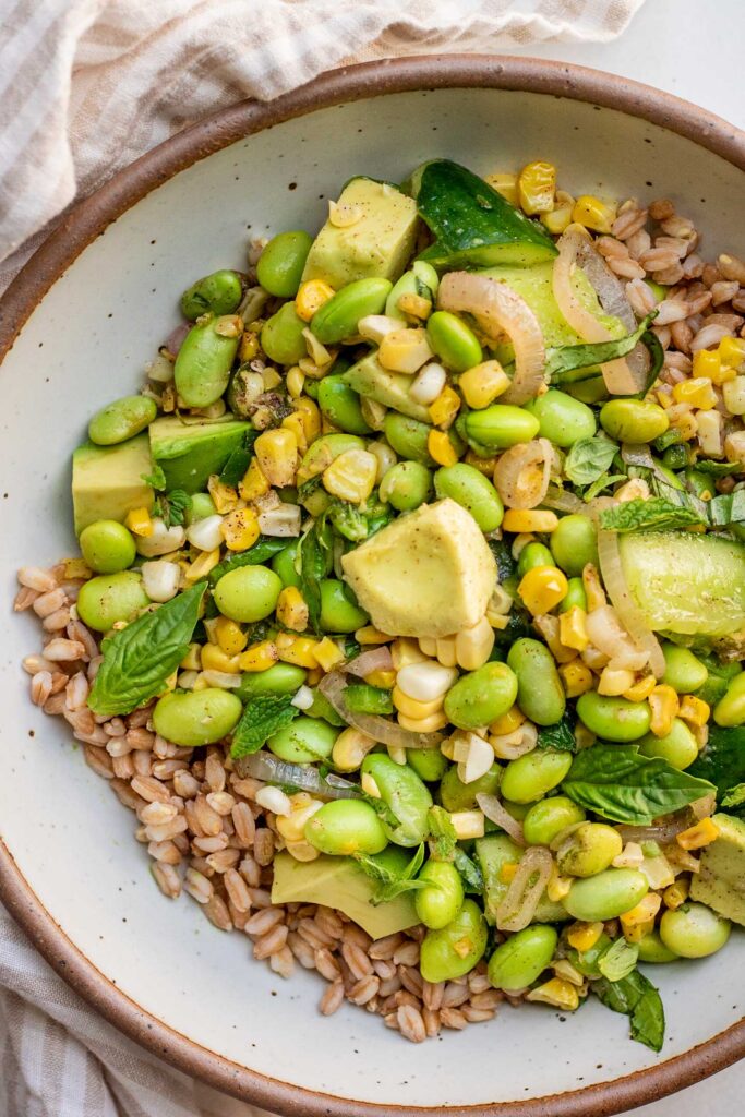 Close up of a bowl of farro topped with cucumber, edamame, corn in a lime sumac vinaigrette.