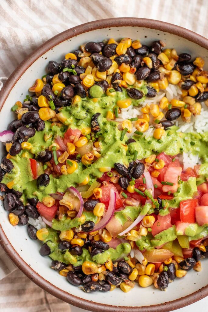 Close up of a rimmed white bowl with rice, black beans and corn topped with tomato salad and a green basil sauce.
