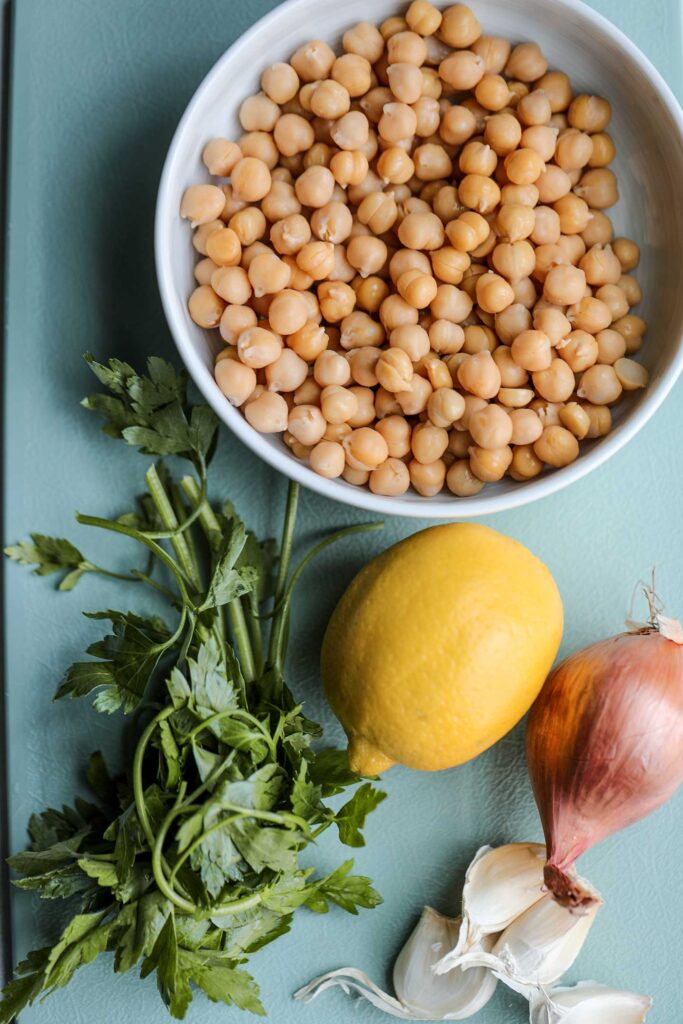 A bowl of chickpeas, lemon, shallot, garlic cloves and bunch of parsley on a cutting board.