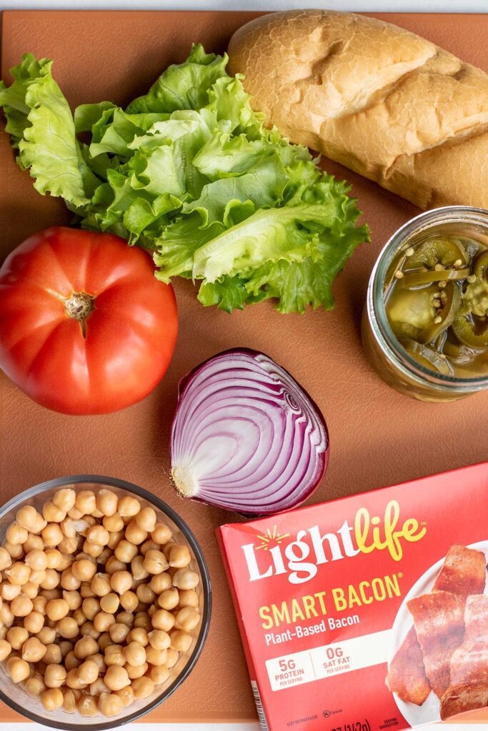 Coral cutting board topped with lettuce, roll, pickled jalapeno, tomatoes, onions, chickpeas and LightLife Smart Bacon box.
