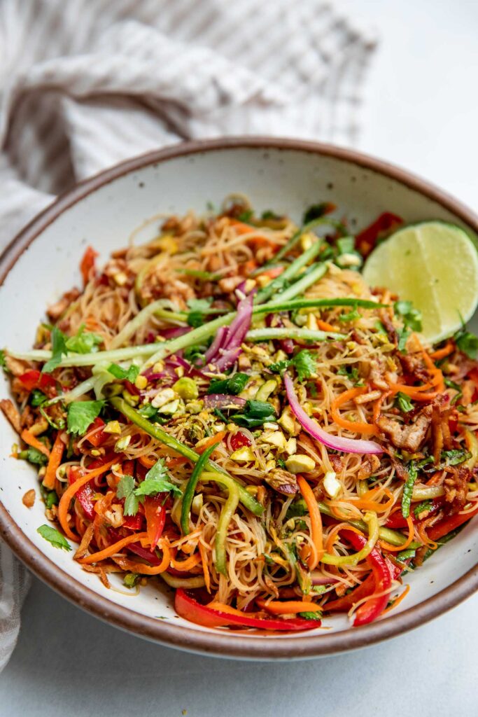 Side view of a bowl of cold noodles mixed with shredded veggies, tofu and maple lime dressing served with a wedge of lime.