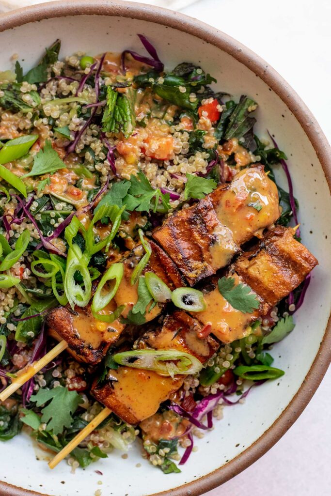 Close up of a bowl of chopped kale salad topped with tofu skewered and almond dressing.