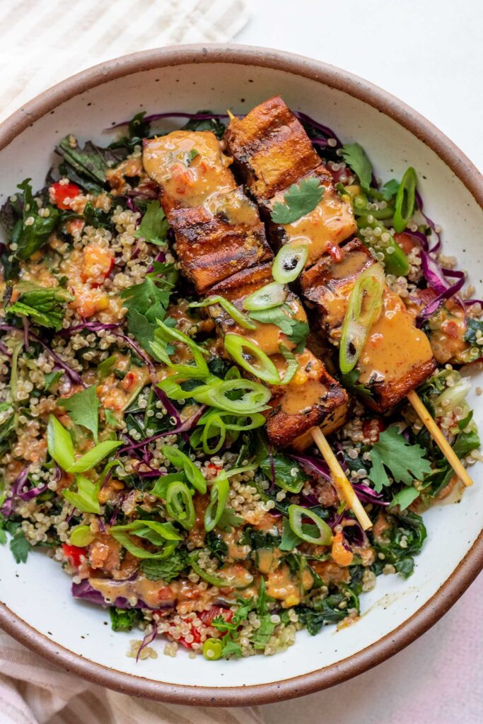 White bowl filled with chopped kale and quinoa salad topped with grilled tofu skewers and almond butter dressing.