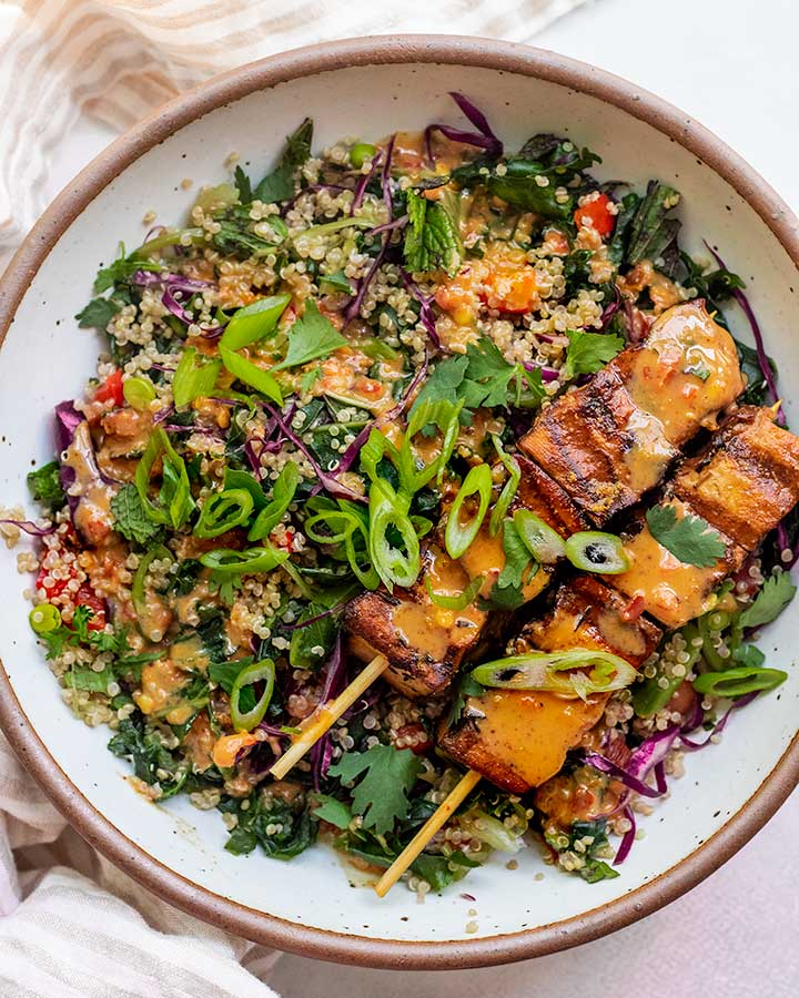 Serving bowl filled with chopped kale quinoa salad and topped with grilled tofu skewers and almond butter dressing.